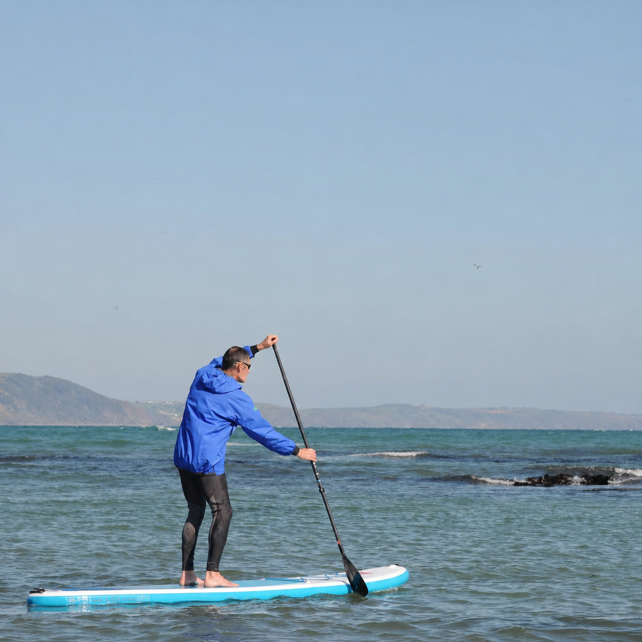 A man stands on a paddleboard on the ocean, wearing a bright blue NKE Centre Smock hooded jacket and Nookie Full Length Wetsuit Strides
